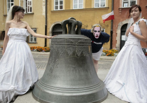 Dziewczyny ubrane w suknie ślubne są po prawej i lewej stronie zdjęcia; po środku stoi dzwon, a zza niego wychyla się postać z maską Putina