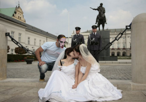 Dwie całujące się i ubrane w suknie ślubne dziewczyny siedzą przed pałacem prezydenckim, za nimi jest dwóch policjantów pełniących wartę, po lewo znów postać z maską Putina patrzy w stronę dziewczyn.