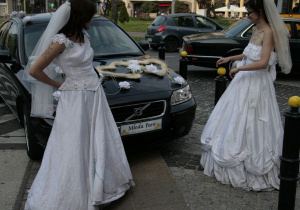Po bokach dziewczyny w sukniach ślubnych pomiędzy nimi bardziej z lewej auto z napisem w miejscu rejestracji 